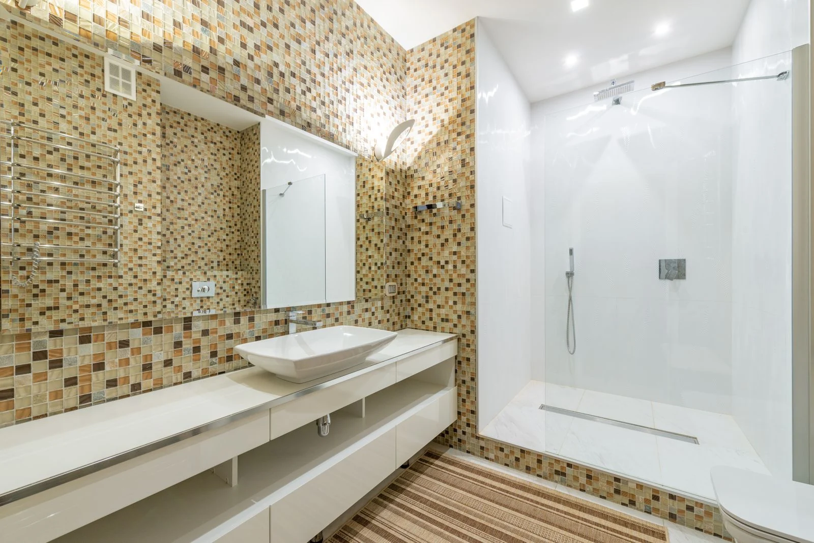 Wide angle of interior of contemporary bathroom with shower cabin and large mirror above white clean sink at mosaic tiled wall